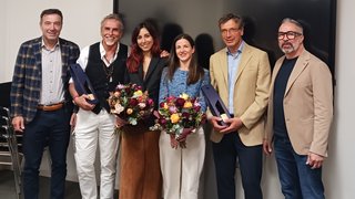 News Sechs Personen stehen zusammen bei einer Feier, zwei halten Blumen und drei Geschenke