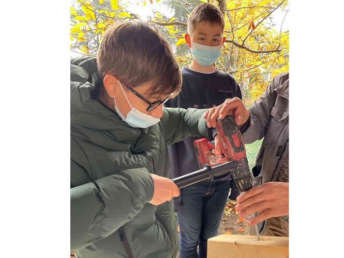 Das „Lebende Handwerk“ in Kaltern Kind benutzt Bohrmaschine unter Anleitung im Freien mit anderen Kindern im Hintergrund