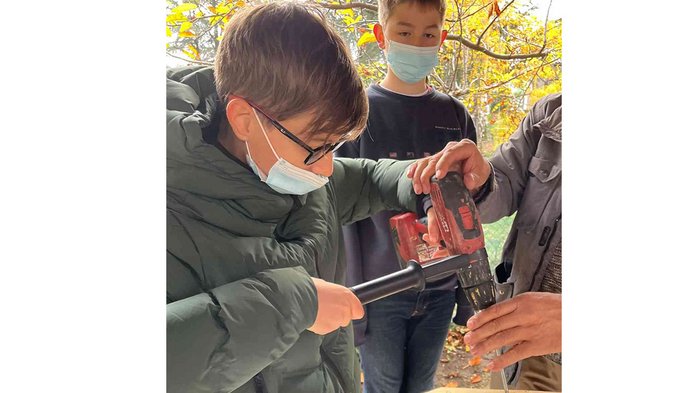 Das „Lebende Handwerk“ in Kaltern Kind benutzt Bohrmaschine unter Anleitung im Freien mit anderen Kindern im Hintergrund