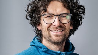 News Lächelnder Mann mit lockigem Haar und Brille in blauem Hoodie vor grauem Hintergrund