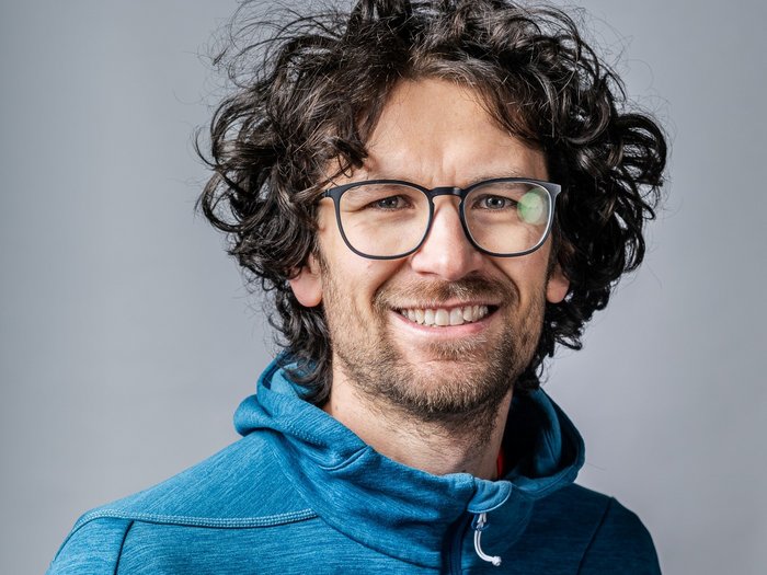 Endlich: Neue staatliche Preisregelung Lächelnder Mann mit lockigem Haar und Brille in blauem Hoodie vor grauem Hintergrund