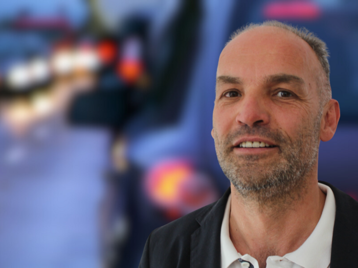 “Evitare un ingorgo colossale” Uomo con giacca nera davanti a sfondo sfocato con veicoli al crepuscolo