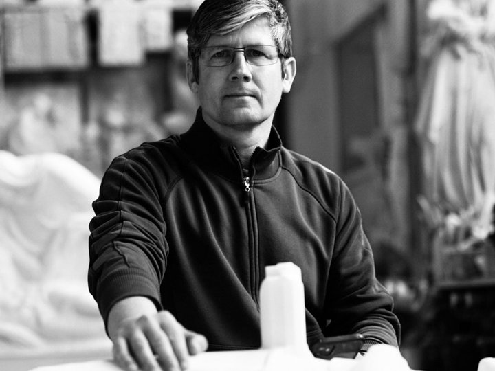 Mayr Josef & Co. s.a.s. Uomo con occhiali e maglione seduto a un tavolo con sculture sullo sfondo