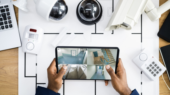 Sicherheit im Betrieb: Land fördert Alarm- und Videoanlagen Person hält Tablet mit Überwachungskamera-Feeds über einem Tisch mit Sicherheitsausrüstung