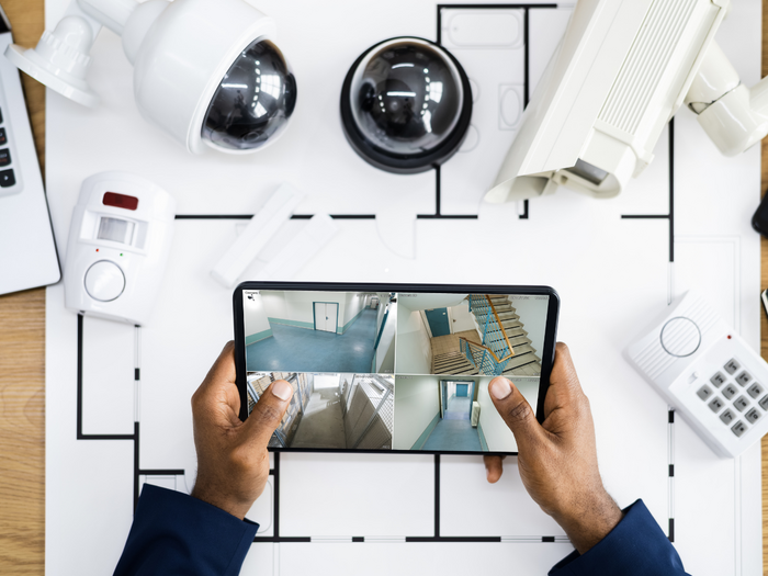 Sicherheit im Betrieb: Land fördert Alarm- und Videoanlagen Person hält Tablet mit Überwachungskamera-Feeds über einem Tisch mit Sicherheitsausrüstung