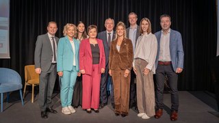 News Neun Personen in Businesskleidung posieren vor schwarzem Vorhang auf Bühne