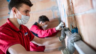 News Zwei Handwerker mit Masken, die an einer Wand in einem Rohbau arbeiten