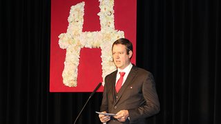 News Mann hält Rede vor rotem Blumen-H auf schwarzem Hintergrund