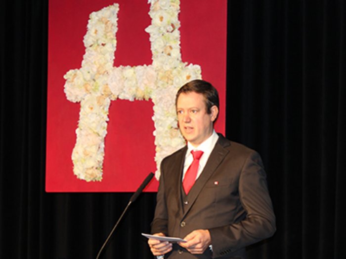 „Kinder, werdet Handwerker!“ Mann hält Rede vor rotem Blumen-H auf schwarzem Hintergrund