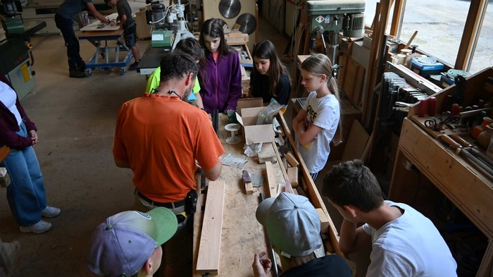 Seifenkistenrennen in St. Felix: Handwerk im Rausch der Geschwindigkeit Gruppe Kinder beobachtet Mann, der in Werkstatt Holz bearbeitet