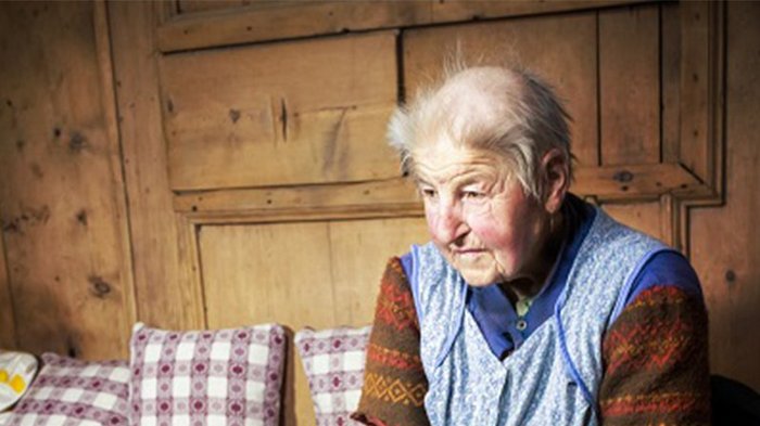 „Nicht den Mut verlieren“ Ältere Frau mit traditioneller Kleidung sitzt auf einem Sofa vor Holzwand