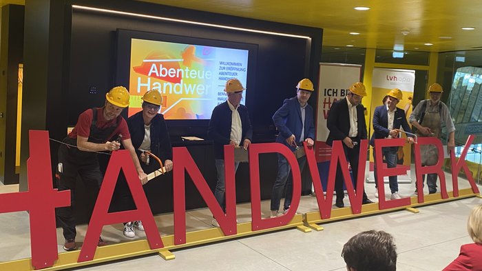 "Abenteuer Handwerk" - Initiative begeistert junge Generation für Handwerksberufe im Pustertal Personen mit Schutzhelmen sägen große rote Buchstaben des Wortes Handwerk