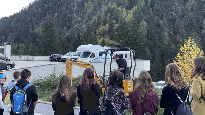 © Mitteschule Pankraz und Ulten Gruppe von Jugendlichen beobachtet einen Mann mit einem gelben Minibagger im Freien vor bewaldetem Berg