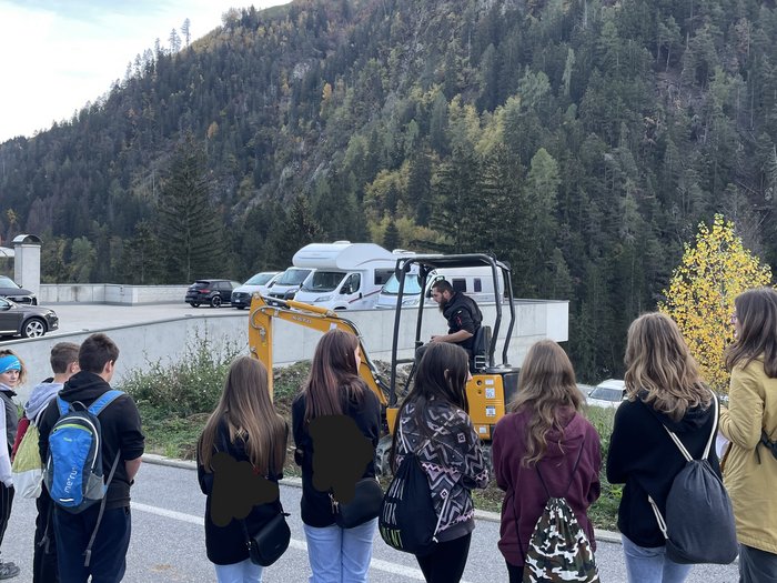 © Mitteschule Pankraz und Ulten Gruppe von Jugendlichen beobachtet einen Mann mit einem gelben Minibagger im Freien vor bewaldetem Berg