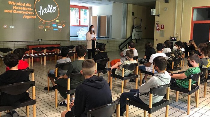 Berufsinfokampagne: Gut informiert in den Sommer Jugendliche sitzen mit Abstand auf Stühlen, Frau mit Maske spricht vor Projektion