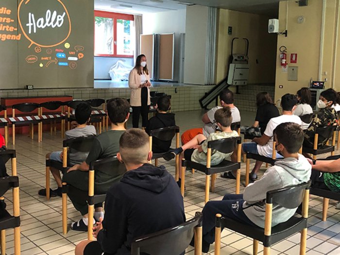 Berufsinfokampagne: Gut informiert in den Sommer Jugendliche sitzen mit Abstand auf Stühlen, Frau mit Maske spricht vor Projektion