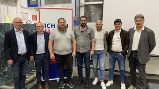 News Sieben Männer in legerer und formeller Kleidung stehen nebeneinander in einer Innenraumhalle