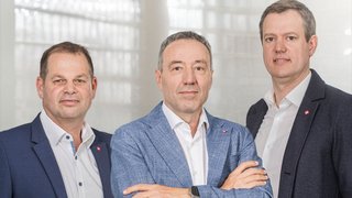 News Drei Männer in Anzügen stehen vor einem hellen Bürofenster