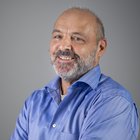 Andreas Baldi Uomo anziano sorridente con barba e camicia blu su sfondo grigio