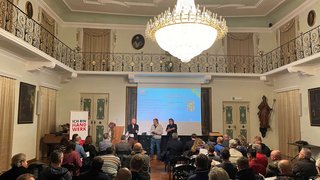 News Vortrag in einem historischen Saal mit großem Kronleuchter und vielen Zuhörern