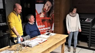 News Drei Personen bei Tisch mit Dokumenten, eine Frau steht und spricht, Werbebanner im Hintergrund