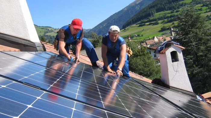 Plattform Land: Energiegemeinschaften – gewusst wie? Zwei Arbeiter installieren Solarpanels auf einem Dach in bergiger Landschaft