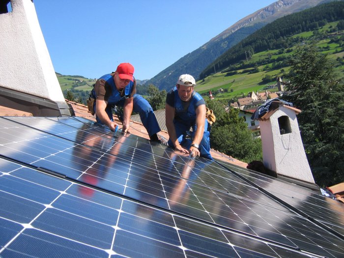 Plattform Land: Energiegemeinschaften – gewusst wie? Zwei Arbeiter installieren Solarpanels auf einem Dach in bergiger Landschaft