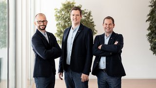 News Drei Männer in Anzügen stehen nebeneinander in einem modernen Büro