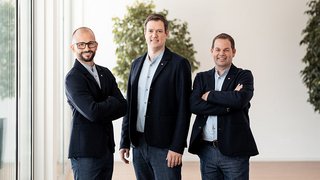 News Drei Männer in Anzügen stehen nebeneinander in einem modernen Büro