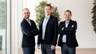 News Drei Männer in Anzügen stehen nebeneinander in einem modernen Büro