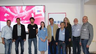 News Neun Personen stehen vor einem Bildschirm und einem Plakat bei einer Veranstaltung.