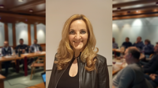 News Lächelnde Frau mit blonden Haaren und schwarzer Jacke vor Gruppenmeeting