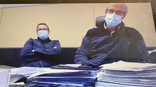 News Zwei Männer mit Masken sitzen hinter einem Tisch mit vielen Akten
