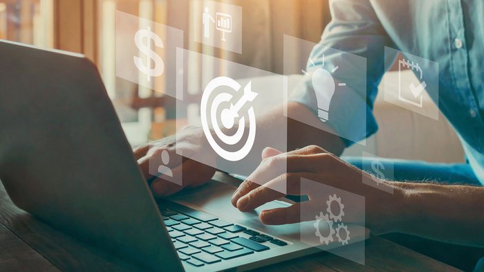 © Song_about_summer/ Shutterstock.com Person tippt auf Laptop mit schwebenden Geschäftssymbolen wie Zielscheibe, Licht und Dollar