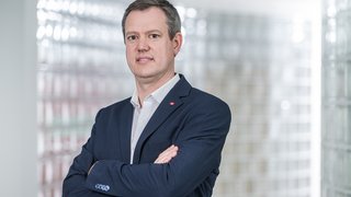 News Mann in dunklem Sakko mit verschränkten Armen vor unscharfem Hintergrund