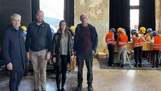 News Vier Erwachsene stehen in einer Halle, im Hintergrund arbeiten Kinder mit Schutzhelmen und Warnwesten