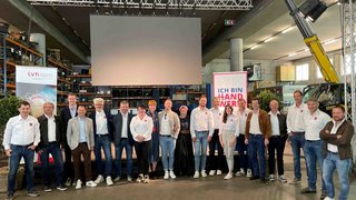 News Gruppenfoto von 18 Personen in einem Lager mit Bannern und Leinwand im Hintergrund