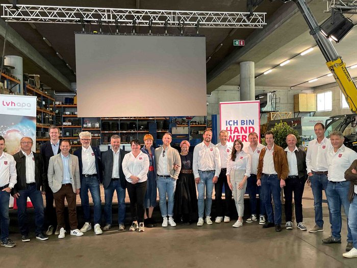 Weltpremiere bei der Jahresversammlung der Baumeister/innen und Maurer/innen Gruppenfoto von 18 Personen in einem Lager mit Bannern und Leinwand im Hintergrund