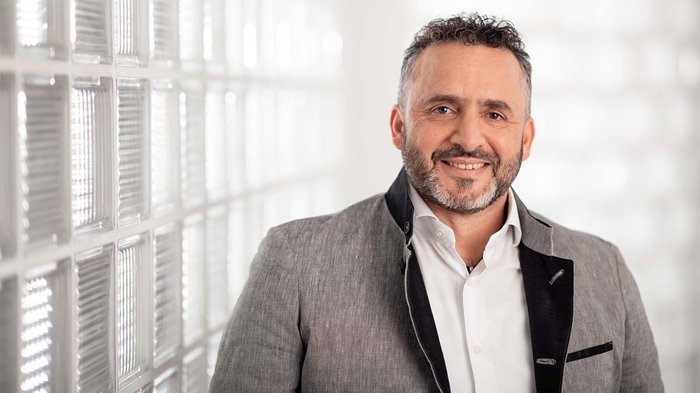 Gruber: „Jetzt klaren Kopf bewahren“ Porträt eines lächelnden Mannes mit grauem Anzug und weißem Hemd vor Glasscheibenwand