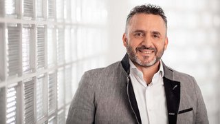 News Porträt eines lächelnden Mannes mit grauem Anzug und weißem Hemd vor Glasscheibenwand