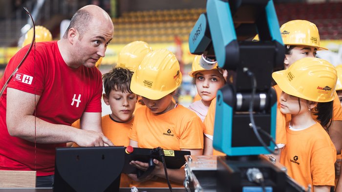 "Avventura artigianato" entusiasma a Merano: i giovani talenti vivono l’artigianato da vicino Adulto spiega a bambini con caschi gialli come usare un robot