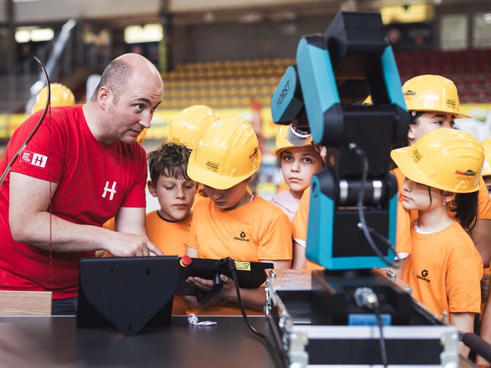 „Abenteuer Handwerk“ begeistert in Meran: Junge Talente erleben das Handwerk hautnah Erwachsener erklärt Kindern in Schutzhelmen Bedienung eines Roboters