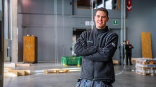 News Giovane uomo con giacca da lavoro grigia sorride in capannone industriale
