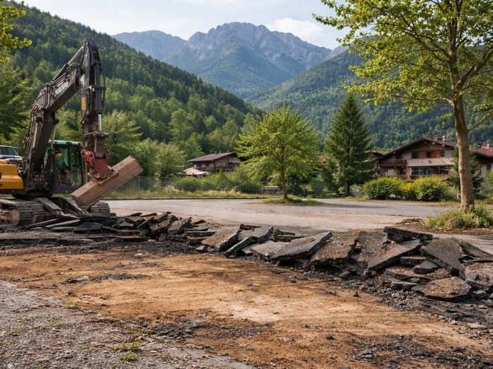 Wettbewerb zur Entsiegelung und Bodenaufwertung 2026 Bagger entfernt Asphalt vor Häusern in einem bewaldeten Tal mit Bergen