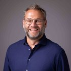 Eduard Zingerle Lächelnder Mann mit Brille und blauem Hemd vor grauem Hintergrund