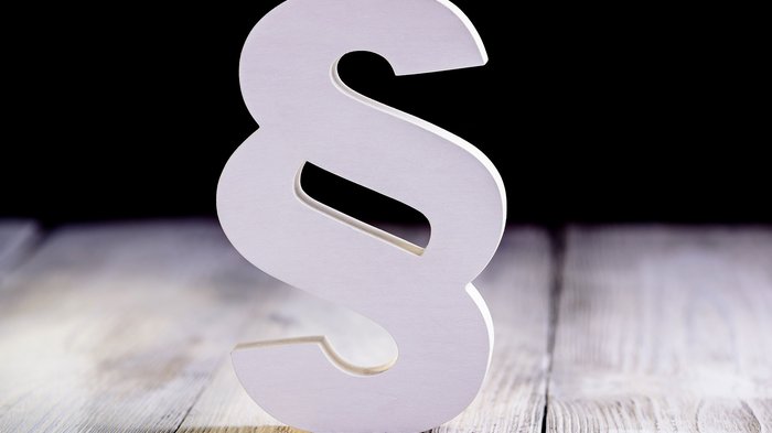 © Daniel Jedzura/Shutterstock.com Weißes Paragraphen-Symbol auf Holzboden vor schwarzem Hintergrund