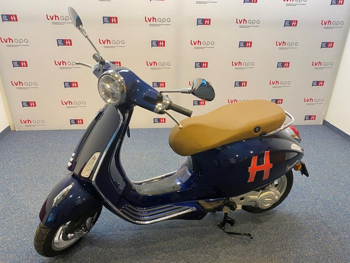 Generation-H-Vespa wird im Vinschgau unterwegs sein Blauer Motorroller mit braunem Sitz vor Werbewand