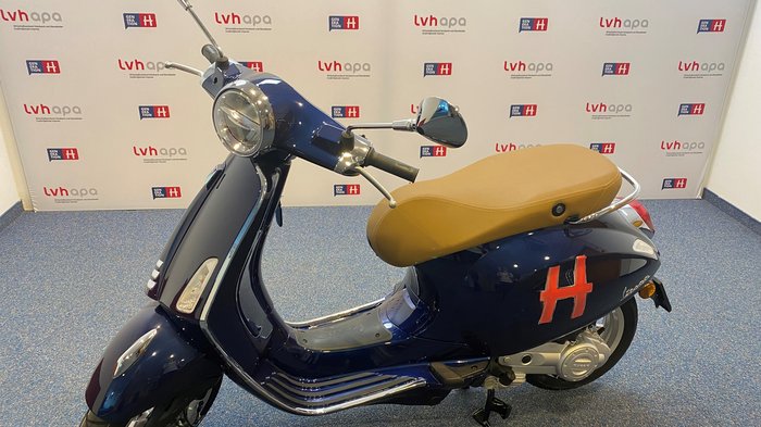 Generation-H-Vespa wird im Vinschgau unterwegs sein Blauer Motorroller mit braunem Sitz vor Werbewand