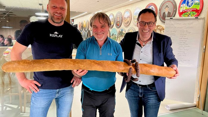 Bezirksschießen der Handwerker/innen Drei Männer halten ein langes Baguette mit Wurst in einem Raum voller Wanddekorationen