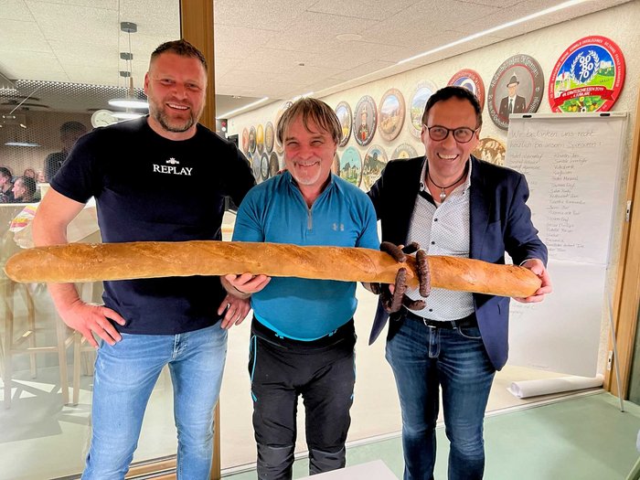 Bezirksschießen der Handwerker/innen Drei Männer halten ein langes Baguette mit Wurst in einem Raum voller Wanddekorationen